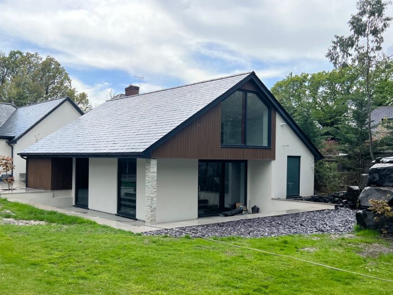 Decking Small raised grey wooden deck in a garden with a round glass-top table and two wicker chairs. The deck sits beside grey patio tiles and is b